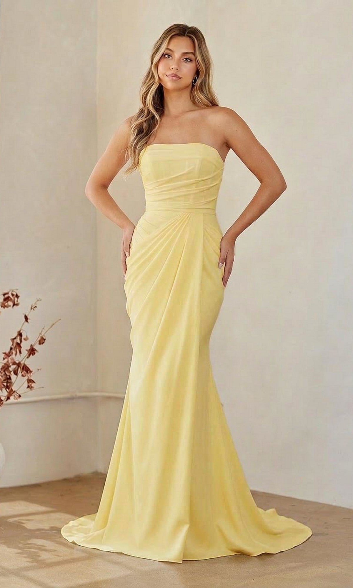 A woman in a yellow Long Formal Dress: Amelia Couture 6149 stands indoors against a neutral background, with dried foliage nearby.
